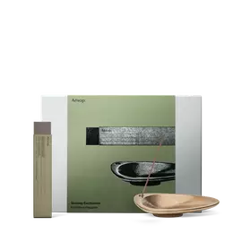 Un baton d’encens repose dans un porte-encens couleur bronze, a cote d’un etui elance, devant un coffret cadeau Aesop vert olive.