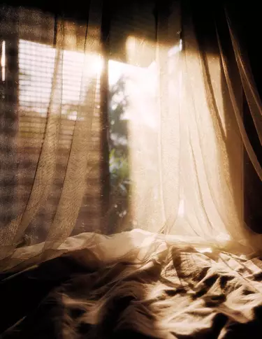 Des rideaux diaphanes drapes sur un tissu froisse pres d’une fenetre baignee de soleil, revelant des tons beige chaud, de douces ombres et des plis translucides.