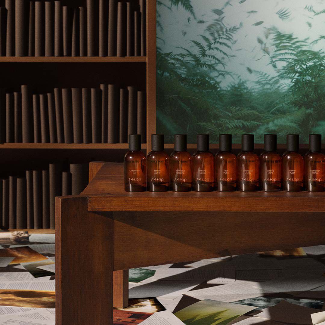 Une rangee de flacons de parfum en verre ambre Aesop exposes sur une etagere en bois devant une bibliotheque et une oeuvre d'art verte.