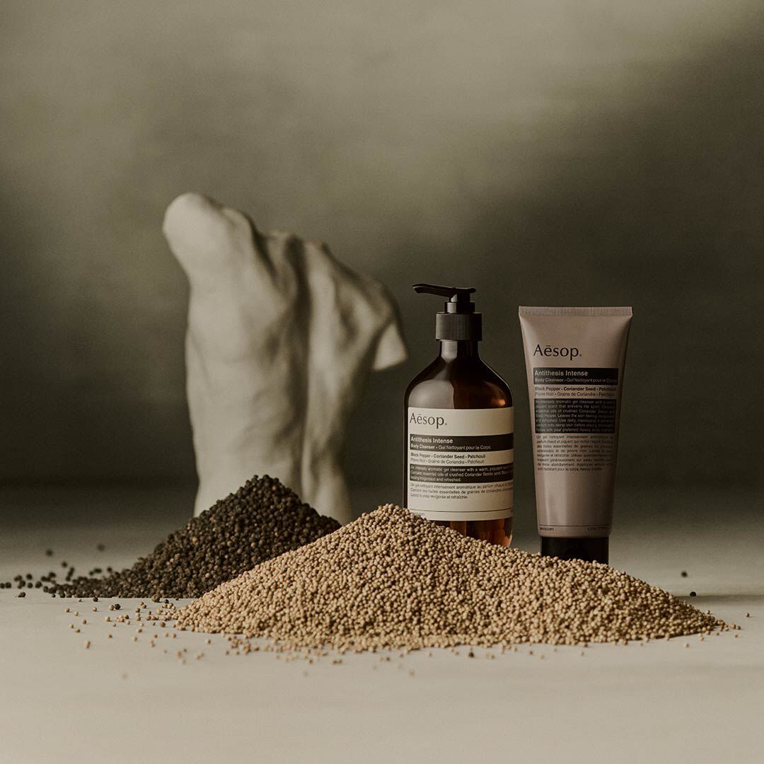 Aesop Antithesis Intense Body Cleanser pump bottle and tube beside piles of black peppercorns and coriander seeds.