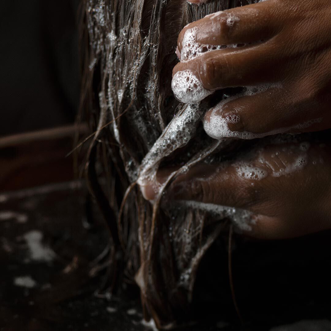 Hands lathering wet hair with soap bubbles over a basin.
