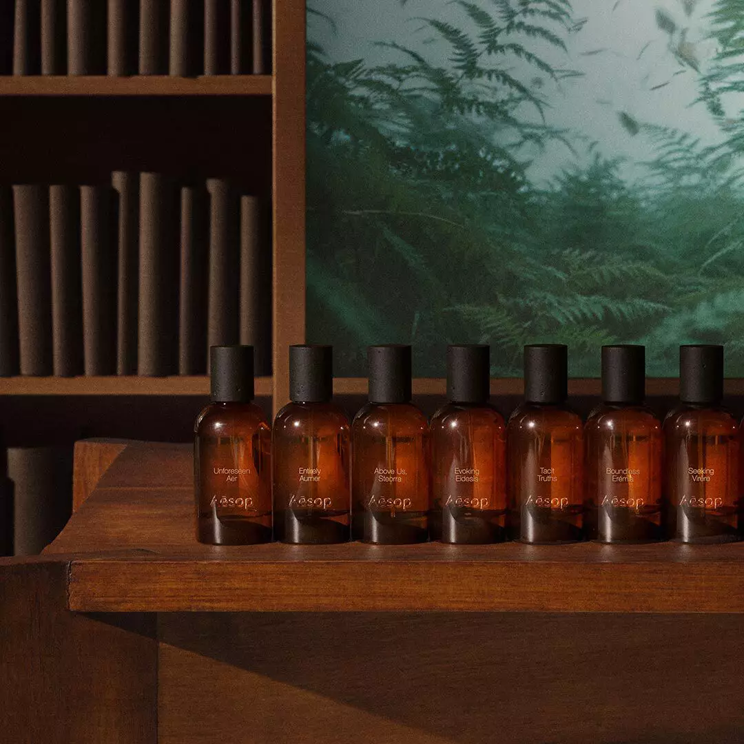 Rangee de flacons de fragrance Aesop ambres sur une table en bois, pres d’etageres et d’une image de fougere verte.