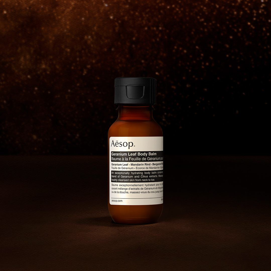 Un flacon de baume pour le corps Geranium Leaf d'Aesop sur un fond sombre et texture.