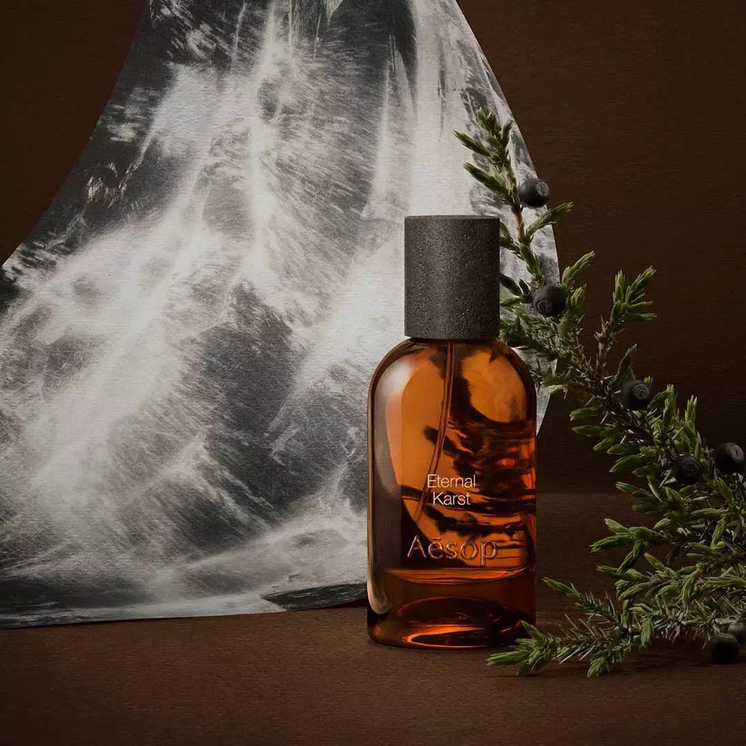 Flacon de fragrance Aesop ambre, pose pres de brins de romarin et d’une image monochrome d’eau, sur une surface brun fonce.