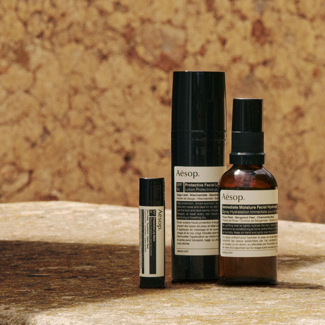 Une selection soignee de formulations de protection solaire Aesop disposees sur une surface en pierre baignee de soleil, contre un mur aux tons chauds de terre.