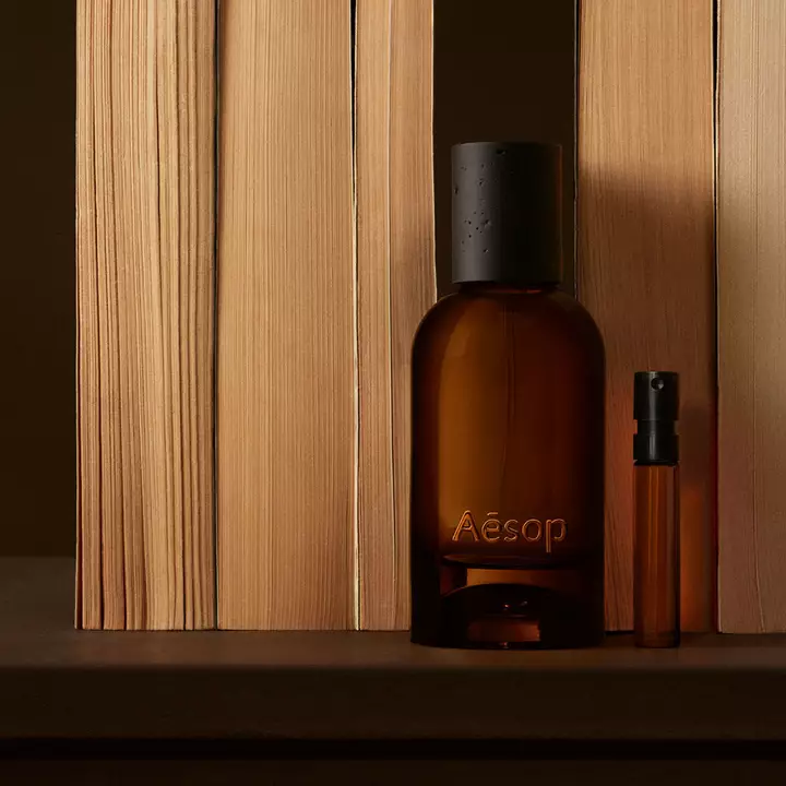Flacon Aesop ambre et fine fiole disposes entre des planches verticales de bois clair, dans de profondes ombres brunes, selon une composition lineaire epuree.