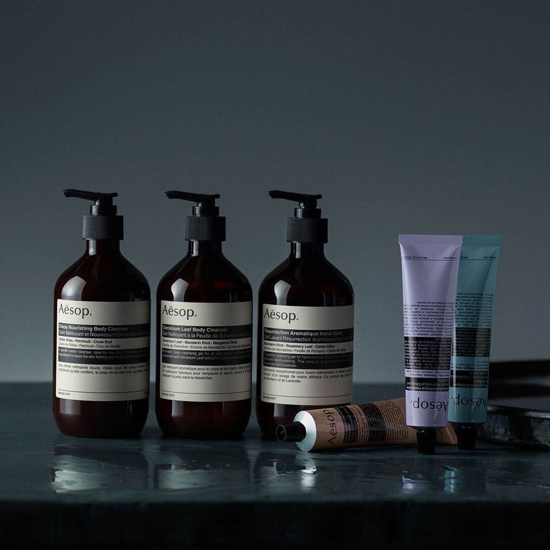 Three Aesop pump bottles, two tubes, and a small amber bottle on a dark reflective surface.