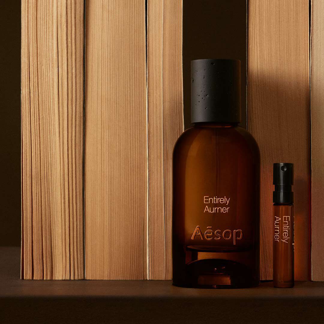 Un flacon en verre ambre de parfum Aesop Entirely Aurner et un echantillon assorti poses verticalement a cote de livres alignes sur une surface sombre.