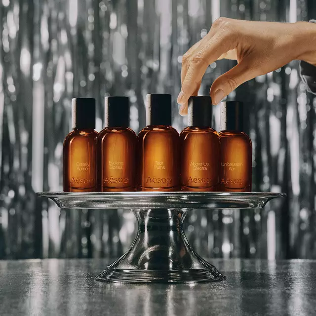 Une rangee de flacons de parfum en verre ambre Aesop exposes sur une etagere en bois devant une bibliotheque et une oeuvre d'art verte.