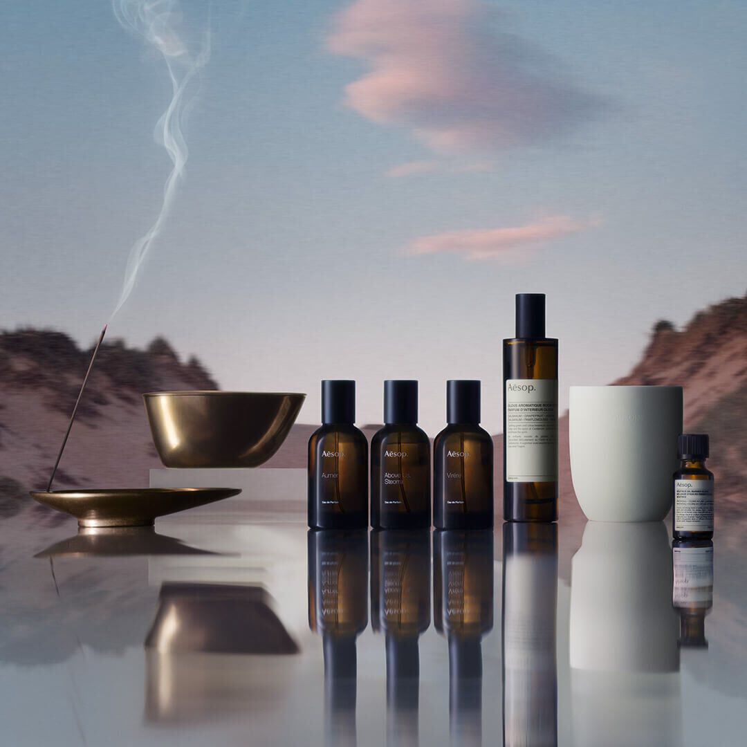 Une collection de produits Aesop, comprenant des parfums, des parfums d'interieur et une huile essentielle, avec un baton d'encens qui brule dans un porte-encens en bronze, pose devant un paysage montagneux flou, reflete sur une surface.