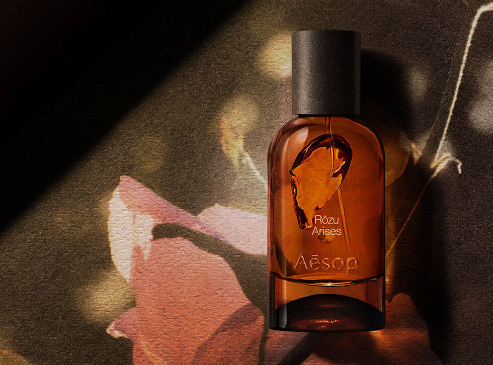 Flacon ambre de fragrance Aesop Rozu Arises sur une surface sombre, a cote d’une illustration botanique rose et d’une racine claire.