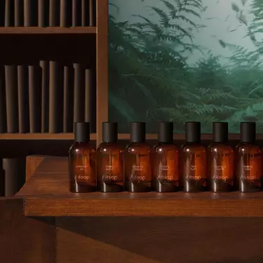 Rangee de flacons de parfum en verre ambre, coiffes de bouchons noirs, sur des etageres en bois a gradins, avec des livres et des fougeres en arriere-plan.